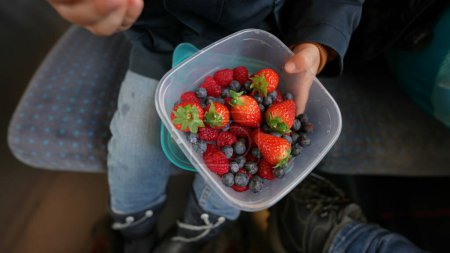 Téléchargez les photos : Gros plan de la main de l'enfant saisissant un conteneur rempli de fraises et de bleuets, assis à bord d'un train - en image libre de droit