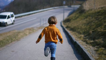 Téléchargez les photos : Par derrière, un enfant fougueux dans un pull jaune, un jean et des bottes sprinte avec délice à travers le paysage automnal. La course animée incarne le frisson de l'air frais de la chute - en image libre de droit