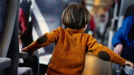 Photo for Young boy in a sunny yellow pullover navigates the train corridor, hands grasping the seat armrests. The little passenger kills time during his rail journey in standout attire - Royalty Free Image