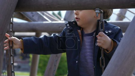 Foto de Niño enfocado jugando en la estructura del patio de recreo, cruzando el puente y sosteniéndose en cadenas de metal de pie en escaleras de madera - Imagen libre de derechos