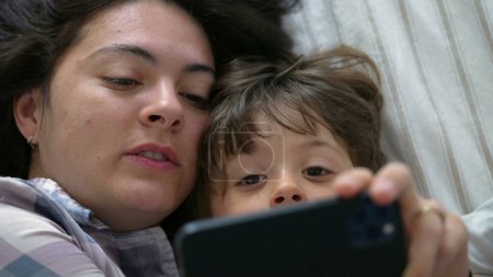 Téléchargez les photos : Enfant et maman engagés dans le lit, concentrés sur l'écran du téléphone comme points d'enfant au contenu en ligne - en image libre de droit