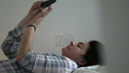 Téléchargez les photos : Femme franche regardant le téléphone posé dans le lit portant un pyjama. Scène domestique décontractée de la personne utilisant un appareil smartphone dans la chambre - en image libre de droit