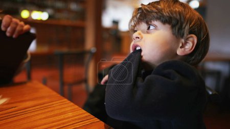 Foto de Niño aburrido tirando de la manga con los dientes y la boca mientras está sentado en el restaurante sin nada que hacer. Niño muerde suéter sensación de aburrimiento - Imagen libre de derechos
