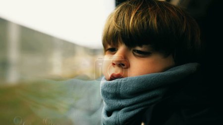Téléchargez les photos : Enfant réfléchi regardant le paysage passer assis par la fenêtre du train appuyé sur la fenêtre portant l'écharpe. Émotion pensive de l'enfant - en image libre de droit