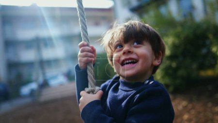 Foto de Acogedor primer plano de niño alegre deslizándose en la cuerda de alambre entre los árboles, Nostalgic Park Fun. Joven sosteniendo fuerte y deslizándose hacia abajo - Imagen libre de derechos