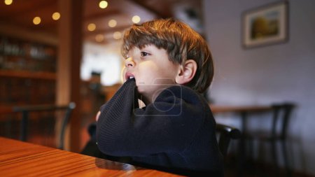 Photo for Bored small boy pulling sleeve with teeth and mouth while seated at restaurant with nothing to do. Child bites sweater feeling boredom - Royalty Free Image