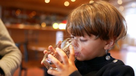 Téléchargez les photos : Petit garçon sirotant de l'eau au restaurant. Enfant buvant et s'hydratant tenant du verre - en image libre de droit
