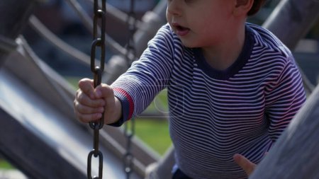 Foto de El niño se apodera de la cadena en la estructura del patio de recreo, perdiendo el equilibrio, aprendiendo a jugar. Niño pequeño en el parque público - Imagen libre de derechos