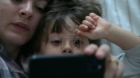 Foto de Niño apuntando a la pantalla del teléfono celular mientras está acostado en la cama junto a la mamá uniéndose mirando el contenido del teléfono en línea - Imagen libre de derechos