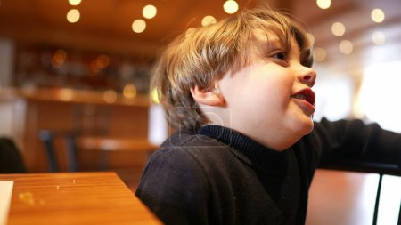 Photo for One small boy sitting at restaurant waiting for food to arrive feeling impatient and idle but with smile and happy - Royalty Free Image