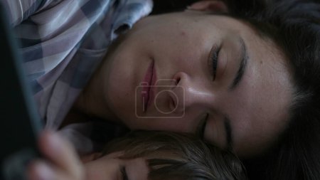 Photo for Close-up of mother and child looking at cellphone device screen laid in bed. Parent and son absorbed by entertainment content online - Royalty Free Image