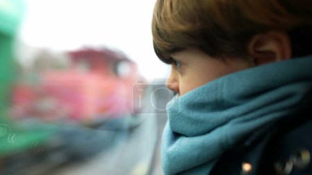 Photo for Little boy leaning on train window looking at landscape pass by at high-speed. Child wearing scarf staring at view in motion - Royalty Free Image