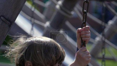 Foto de El niño se apodera de la cadena en la estructura del patio de recreo, perdiendo el equilibrio, aprendiendo a jugar. Niño pequeño en el parque público - Imagen libre de derechos