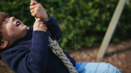 Foto de Feliz cuerda infantil deslizándose en el parque entre dos árboles. Pequeño niño sintiendo emoción durante el otoño día de otoño - Imagen libre de derechos