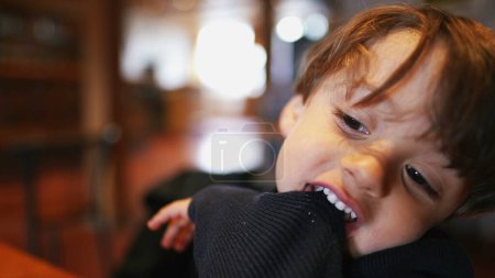Photo for Bored small boy pulling sleeve with teeth and mouth while seated at restaurant with nothing to do. Child bites sweater feeling boredom - Royalty Free Image