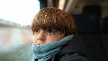 Foto de Niño juguetón viajando en tren apoyado en un cristal de ventana usando una tarjeta, girando la cabeza hacia los lados mientras el paisaje pasa a alta velocidad - Imagen libre de derechos