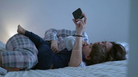 Foto de Madre e hijo viendo contenido en línea acostados en la cama juntos, mamá sosteniendo dispositivo de teléfono inteligente con una mano, escena doméstica sincera y auténtica de los padres y el hijo - Imagen libre de derechos