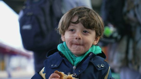 Foto de Retrato de un niño feliz deleitado comiendo croissant de pie en la plataforma del tren con chaqueta y bufanda durante la temporada de otoño otoño. Comida de carbohidratos para niños con una amplia sonrisa - Imagen libre de derechos