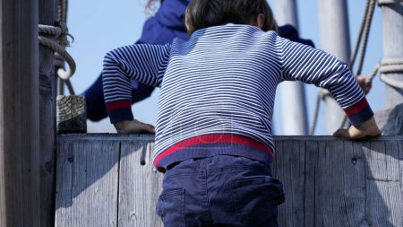 Téléchargez les photos : Petit garçon escalade structure de terrain de jeu maintenant l'équilibre. Enfant montant sur une échelle en bois - en image libre de droit