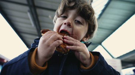 Téléchargez les photos : Portrait d'enfant enchanté avec un large sourire, mangeant croissant à la plate-forme de train dans les vêtements d'automne - en image libre de droit