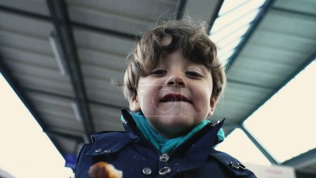 Foto de Retrato de niño encantado con amplia sonrisa, comiendo croissant en la plataforma del tren en ropa de otoño - Imagen libre de derechos