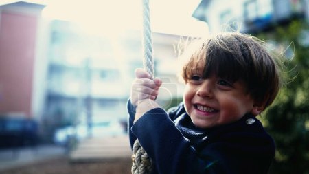 Foto de Niño pequeño alegremente deslizándose por la cuerda de alambre entre árboles en Autumn Park. Alegre niño firmemente agarre y descienda la diapositiva de cuerda de alambre en el parque público en otoño - Imagen libre de derechos