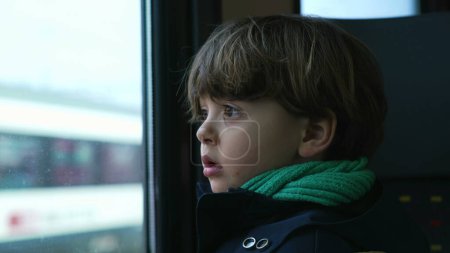 Téléchargez les photos : Introspectif enfant coûteux voyageant en train regardant le paysage passer de la fenêtre de transport portant écharpe et veste pendant la saison froide - en image libre de droit
