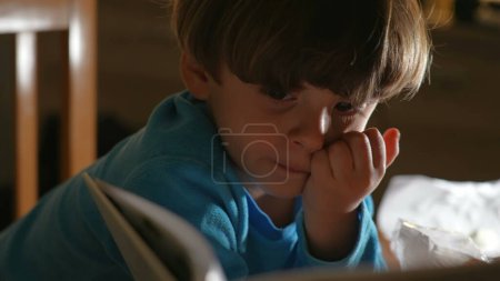 Foto de Un niño pequeño absorbido por el libro de cuentos en la noche antes de acostarse. Primer plano niño mirando el libro, volteando la página - Imagen libre de derechos