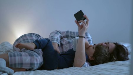 Téléchargez les photos : Mère et enfant regardant du contenu en ligne couché ensemble, maman tenant un appareil smartphone avec une main, scène domestique franche et authentique de parent et fils - en image libre de droit
