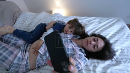Foto de Padres e hijos usando pijamas, acostados en la cama y viendo contenido en el teléfono juntos - Imagen libre de derechos