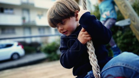 Foto de Niño pequeño alegremente deslizándose por la cuerda de alambre entre árboles en Autumn Park. Alegre niño firmemente agarre y descienda la diapositiva de cuerda de alambre en el parque público en otoño - Imagen libre de derechos