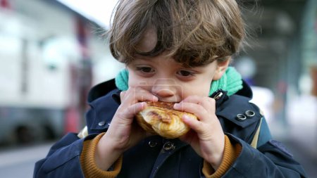 Photo for Happy small boy eating croissant standing at train platform wearing jacket and warm clothes during fall autumn season. Portrait of kid snacking carb food - Royalty Free Image