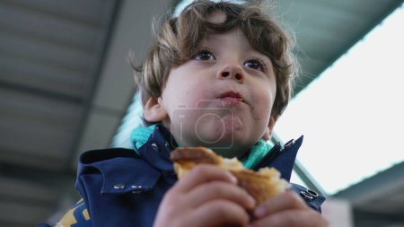 Foto de Joyful Kid merienda en Croissant en la estación de tren, vestido con chaqueta de otoño y bufanda - Imagen libre de derechos