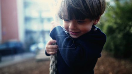 Téléchargez les photos : Petit garçon glissant joyeusement le long de la corde métallique entre les arbres dans Autumn Park. Joyeux enfant fermement accrocheur et descendant fil glissement de corde au parc public à l'automne - en image libre de droit