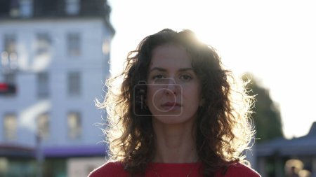 Téléchargez les photos : Portrait d'une jeune femme confiante debout à l'extérieur dans la rue de la ville pendant le coucher du soleil regardant directement la caméra rétroéclairée - en image libre de droit