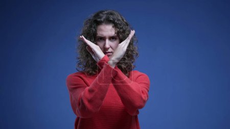 Photo for Young woman saying NO to camera by pointing and waving finger. 20s female person making cross with arms rejecting offer or behavior standing on blue backdrop - Royalty Free Image
