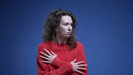 Foto de Mujer joven asustada abrazándose vacilante mostrando miedo y entrando en pánico sobre fondo azul usando suéter rojo. veinteañera mujer miedo - Imagen libre de derechos