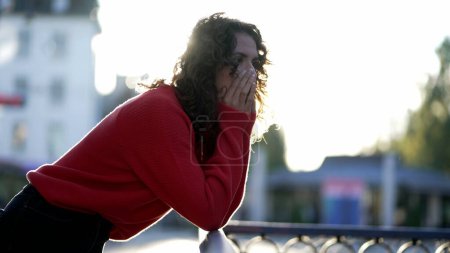 Foto de Mujer reflexiva contemplando la distancia al atardecer de la ciudad apoyada en un riel metálico con destellos de luz solar brillando en el fondo. 20s persona femenina en profunda contemplación - Imagen libre de derechos