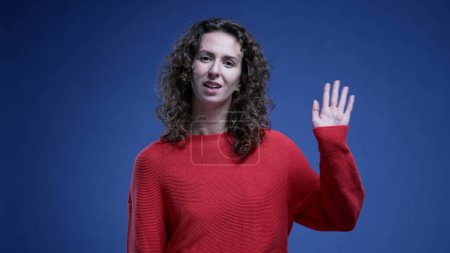 Photo for Happy woman waving hello to camera with hand while standing on a blue background with vibrant red sweater - Royalty Free Image