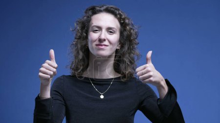 Photo for Woman giving thumbs up on blue background. 20s female person showing approval, positive gesture - Royalty Free Image
