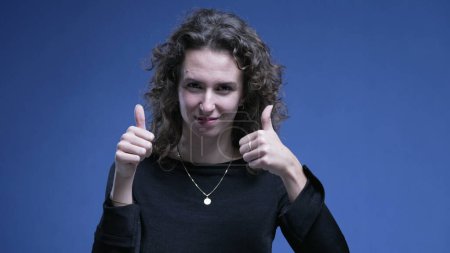 Photo for Woman giving thumbs up on blue background. 20s female person showing approval, positive gesture - Royalty Free Image