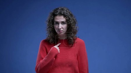 Photo for Incredulous woman flabbergasted by accusation, pointing to herself with finger in disbelief standing on blue backdrop. Funny surprised 20s person points with hand to self - Royalty Free Image