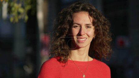 Foto de Mujer feliz de 20 años en la cámara de pie fuera en el entorno urbano de la ciudad durante el atardecer con sol brillando en la cara sonriendo - Imagen libre de derechos