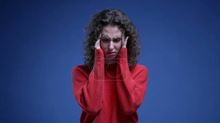 Photo for Anxious young woman with holding head temples with hands struggling with mental anxiety feeling overwhelmed with problems standing on blue background - Royalty Free Image