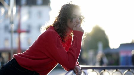 Foto de Mujer joven pensativa apoyada en riel de metal fuera en la calle de la ciudad durante la puesta del sol con la luz del sol resplandor. Expresión reflexiva de la persona con la mano en la barbilla en la contemplación profunda - Imagen libre de derechos