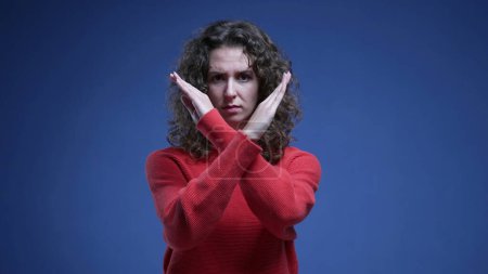 Photo for Young woman saying NO to camera by pointing and waving finger. 20s female person making cross with arms rejecting offer or behavior standing on blue backdrop - Royalty Free Image