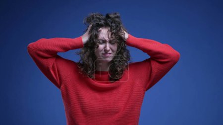 Foto de Mujer joven frustrada tirando de su cabello en la ansiedad y la desesperación en el telón de fondo azul con suéter rojo. Ansiosa veinteañera persona femenina - Imagen libre de derechos