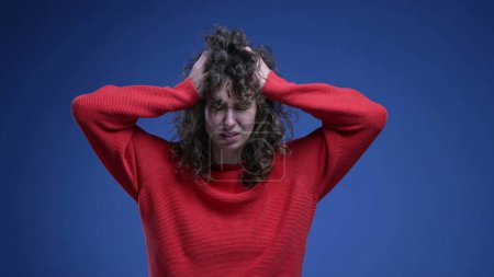 Foto de Mujer joven frustrada tirando de su cabello en la ansiedad y la desesperación en el telón de fondo azul con suéter rojo. Ansiosa veinteañera persona femenina - Imagen libre de derechos