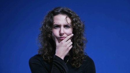 Photo for Pensive woman pondering decision with hand in chin thinking deeply about dilemma. 20s female person with thoughtful expression standing on blue background - Royalty Free Image