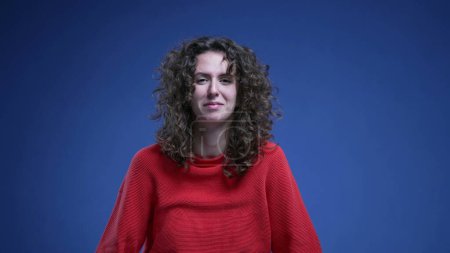 Photo for Happy Woman shrugging dismissing by saying "whatever" with her body language standing on blue backdrop with red sweater - Royalty Free Image